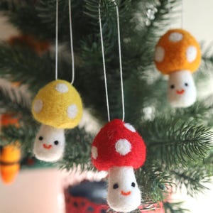 May include: Three felt mushroom ornaments hanging on a Christmas tree. The mushrooms are red with white dots, yellow with white dots, and orange with white dots. Each mushroom has a white stem with a smiling face.