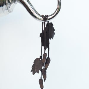 Puede incluir: Un gancho decorativo plateado con un diseño curvo, con un patrón floral detallado en la base. Del gancho cuelgan adornos de hojas y bellotas de color marrón oscuro, suspendidos por finos cordones. El fondo es blanco liso.