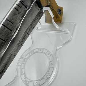 May include: A detailed view of a sword blade and hilt, resting on a clear acrylic stand. The blade has a textured, weathered appearance. The hilt is gold-colored with blue accents. The stand has a circular design.