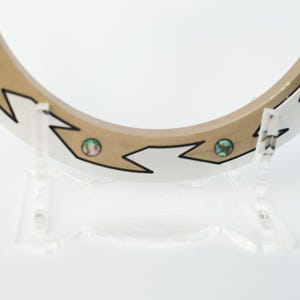 May include: A decorative wooden ring with a light brown center and a white outer edge. The ring features a zigzag design with black outlines and inlaid iridescent accents. It is displayed on a clear acrylic stand.