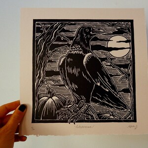 May include: Black and white woodcut print of a raven perched on a branch with pumpkins in the foreground. The raven is facing right and has a large beak. The moon is visible in the background. The print is signed "Redeemer" and dated "12/21".