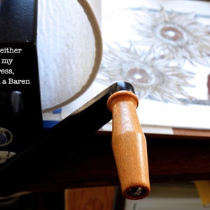May include: A black printing press with a wooden handle. The press is used to create prints by pressing a block of wood or metal onto paper. The text on the press reads: "Each print is either pressed on my table top press, or by hand with a Baren."