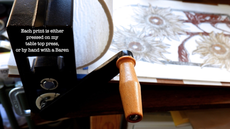 May include: A black printing press with a wooden handle. The press is used to create prints by pressing a block of wood or metal onto paper. The text on the press reads: "Each print is either pressed on my table top press, or by hand with a Baren."