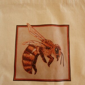 May include: A beige tote bag with a brown frame around a drawing of a bee. The bee is brown and yellow with a large eye. The text "HANNAH JAY ARTWORK" is printed at the top of the bag.