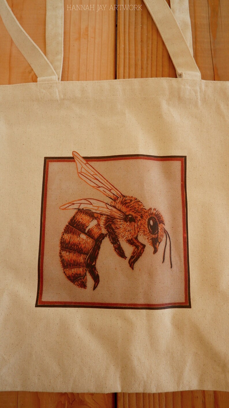 May include: A beige tote bag with a brown frame around a drawing of a bee. The bee is brown and yellow with a large eye. The text "HANNAH JAY ARTWORK" is printed at the top of the bag.