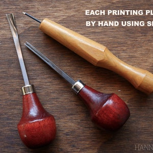 May include: Three wood carving tools with red and brown handles and metal blades. The tools are on a wooden surface. The text "EACH PRINTING PLATE IS CARVED BY HAND USING SMALL CHISELS" is above the tools. The text "HANNAH JAY ARTWORK" is below the tools.