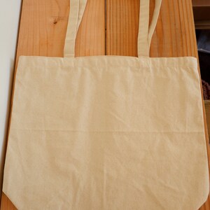 May include: A plain, beige canvas tote bag with two long handles. The bag is lying flat on a wooden surface.