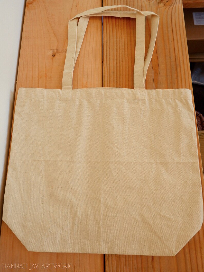 May include: A plain, beige canvas tote bag with two long handles. The bag is lying flat on a wooden surface.