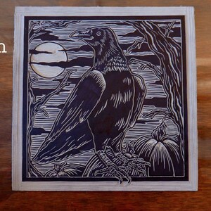 May include: A black and white linoleum block print of a raven perched on a pumpkin with a full moon in the background. The print is titled "Carved Linoleum Plate".