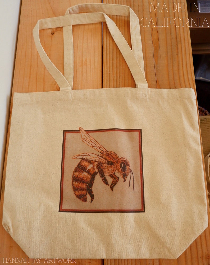 May include: A beige canvas tote bag with brown straps. The bag has a brown square design with a drawing of a bee inside. The text "MADE IN CALIFORNIA" is printed on the top of the bag. The text "HANNAH JAY ARTWORK" is printed on the bottom of the bag.