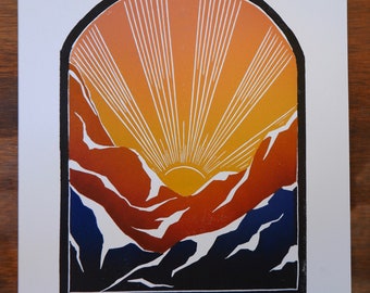 May include: A print featuring a stylized mountain landscape with a rising sun. The artwork uses a palette of orange, yellow, blue, and white, framed by a black arch. The sun's rays burst from behind the mountains.