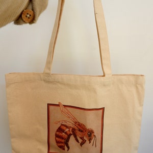 May include: A beige tote bag with a brown bee illustration on the front. The bag is made of 100% cotton.
