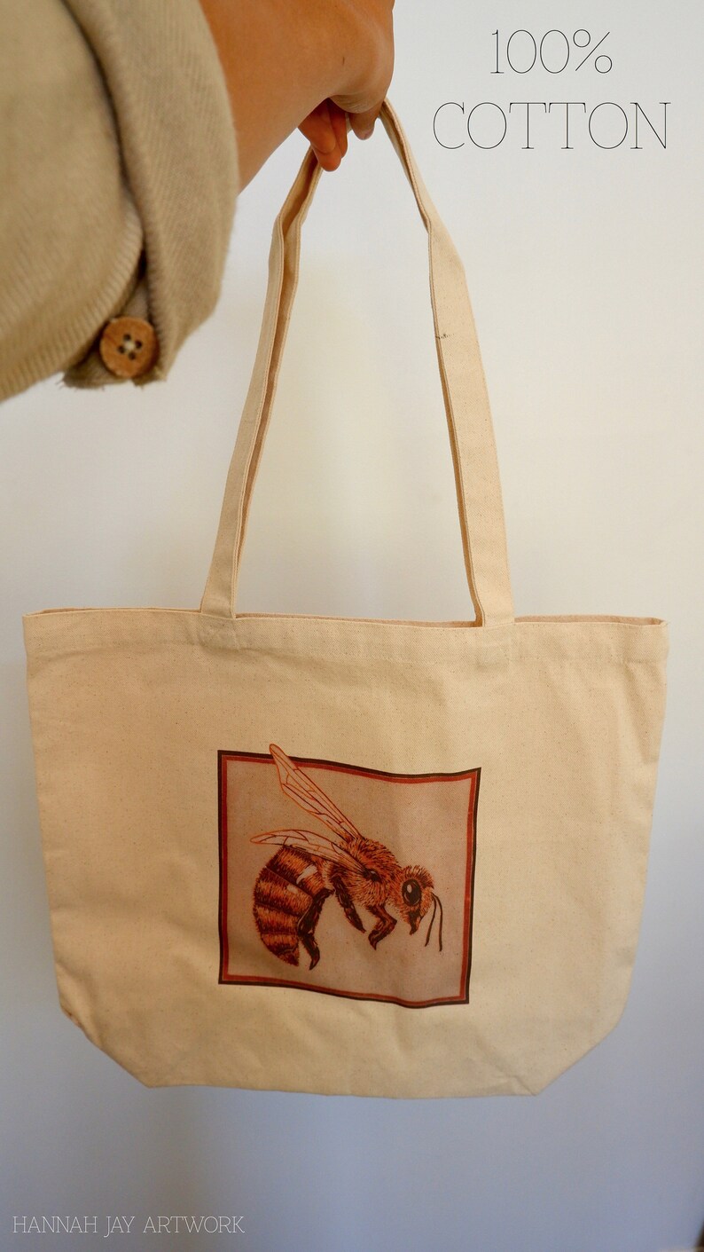 May include: A beige tote bag with a brown bee illustration on the front. The bag is made of 100% cotton.