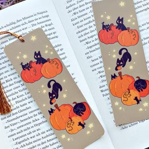 Op de afbeelding: Twee beige bladwijzers met een Halloween-thema liggen op een open boek. Elke bladwijzer heeft illustraties van zwarte katten, oranje pompoenen en kleine sterren. Een bruine kwast hangt aan de bovenkant van een bladwijzer.