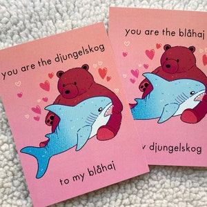 May include: Two pink greeting cards with a cartoon bear hugging a shark. The text reads "You are the djungelskog to my blåhaj" and "You are the blåhaj v djungelskog" with heart illustrations.
