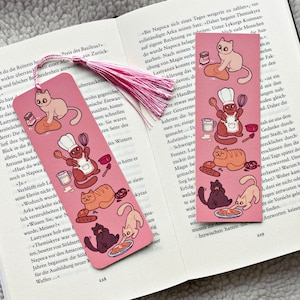 May include: Pink bookmark with a pink tassel featuring illustrations of cats in various poses, including a cat wearing a chef's hat and apron, a cat baking bread, and a cat holding a rolling pin. The bookmark also features a variety of other illustrations, including a loaf of bread, a bowl of flour, and a rolling pin.