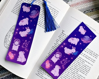 May include: Two purple and blue space-themed bookmarks featuring illustrations of rabbits, planets, and constellations. Each bookmark has a blue tassel. The bookmarks are on an open book.