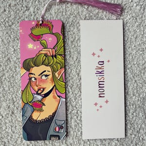 Pu&ograve; includere: Un segnalibro rettangolare con sfondo rosa che presenta l'illustrazione di un personaggio dai capelli verdi e orecchie a punta, circondato da piante carnivore. Il retro &egrave; bianco con il testo "nomsikka" e piccole icone a forma di stella.