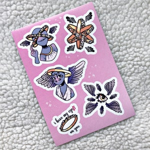May include: A set of four stickers featuring a celestial theme with a purple and blue color scheme. The stickers feature a figure with multiple eyes and wings, a halo with multiple eyes, a ring with multiple eyes, and a single eye with wings. The text "I have my eyes on you" is written on the sticker sheet.
