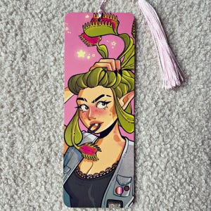 Pu&ograve; includere: Un segnalibro rettangolare con un'illustrazione colorata di un personaggio con capelli verdi acconciati come piante dionee, che indossa un top nero e un gilet grigio con spille, su sfondo rosa con una nappa rosa chiaro attaccata nella parte superiore.
