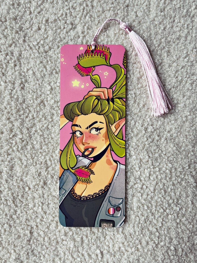 Pu&ograve; includere: Un segnalibro rettangolare con un'illustrazione colorata di un personaggio con capelli verdi acconciati come piante dionee, che indossa un top nero e un gilet grigio con spille, su sfondo rosa con una nappa rosa chiaro attaccata nella parte superiore.
