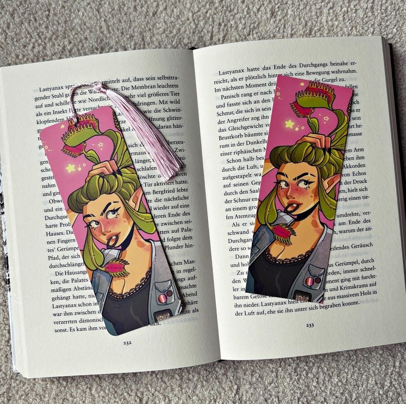 Pu&ograve; includere: Due segnalibri rettangolari con un'illustrazione colorata di un personaggio con capelli verdi acconciati come una dionea acchiappamosche, che indossa un top nero e un gilet grigio. Il segnalibro a sinistra ha una nappa rosa chiaro.