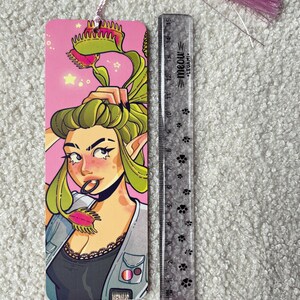 Pu&ograve; includere: Un segnalibro rettangolare con l'illustrazione di un personaggio dai capelli verdi e piante carnivore, completato da una nappa rosa. Accanto si trova un righello in plastica trasparente da 15 cm con impronte nere e la scritta meow.