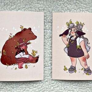 May include: Two illustrated cards with a whimsical theme. The first card features a brown bear with a person wearing a hat and sitting on the bear's back. The second card features a person with grey hair and a white shirt, wearing a purple overall dress with a floral pattern. Two black birds are perched on the person's arms.