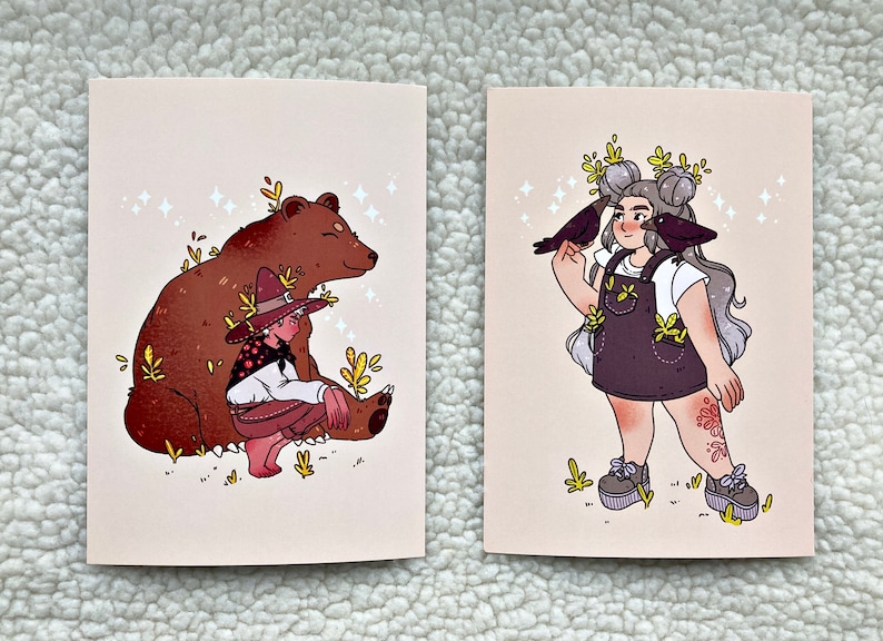 May include: Two illustrated cards with a whimsical theme. The first card features a brown bear with a person wearing a hat and sitting on the bear's back. The second card features a person with grey hair and a white shirt, wearing a purple overall dress with a floral pattern. Two black birds are perched on the person's arms.