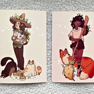 May include: Two illustrated cards featuring women with animal companions. The first card shows a woman with brown hair wearing a brown hat with green leaves and holding two white cats. She is wearing a brown sweater and brown pants. She is standing next to a black cat and a brown and white cat. The second card shows a woman with brown skin and curly black hair wearing a white shirt and black shorts. She is holding a brown broom and is standing next to a red fox.
