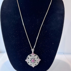 May include: A silver-toned necklace featuring a circular pendant with an intricate openwork filigree design. The pendant is accented with small, sparkling clear stones and several round, vibrant pink gemstones set throughout the metal frame.