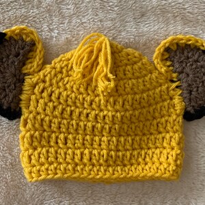 May include: A yellow crocheted hat with brown ears and a yellow tassel. The hat is designed to look like a lion.