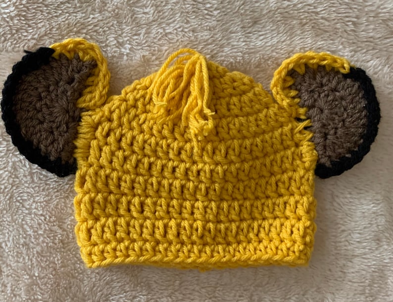 May include: A yellow crocheted hat with brown ears and a yellow tassel. The hat is designed to look like a lion.