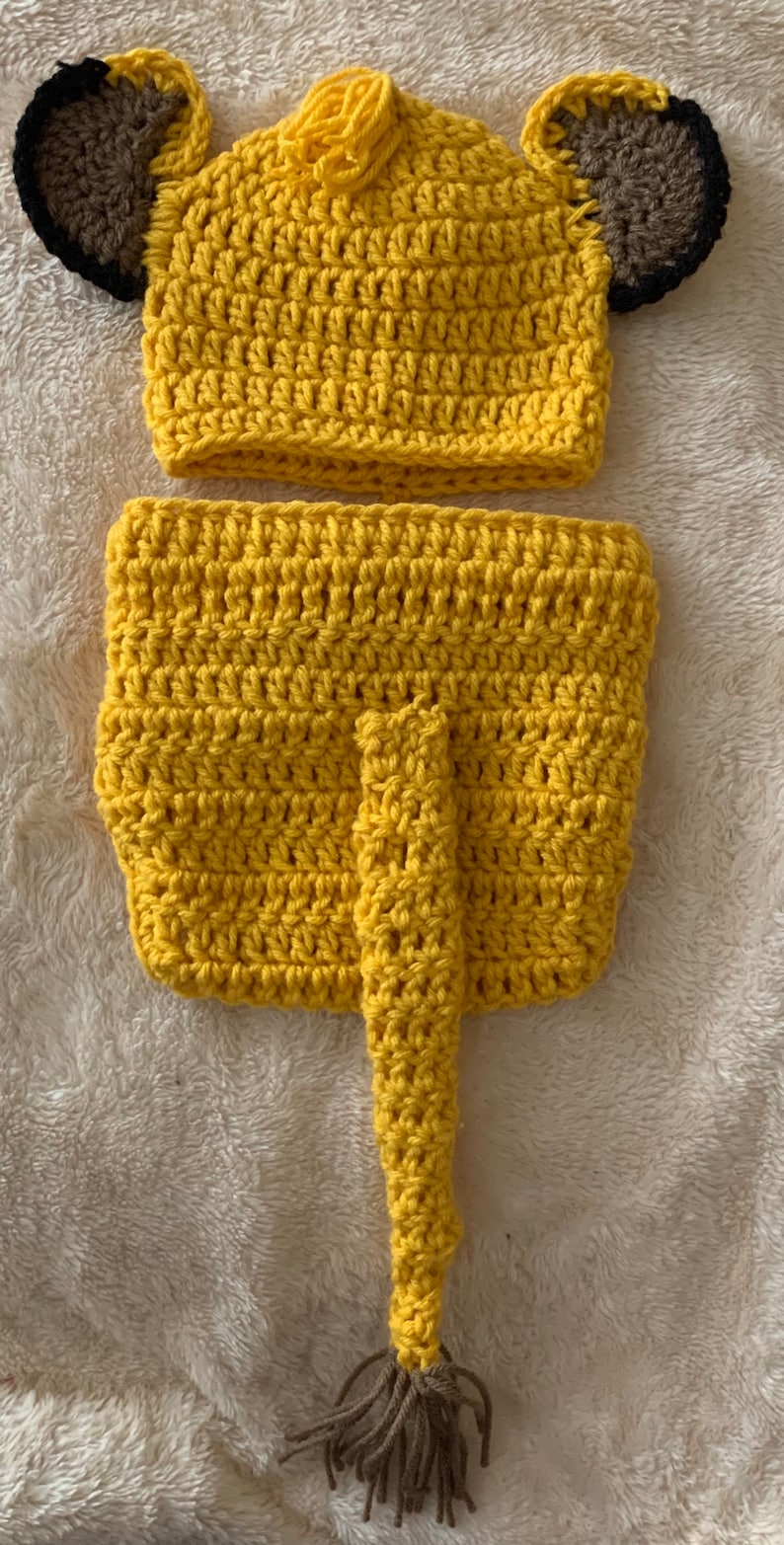 May include: A yellow crocheted lion costume for a baby, featuring a hat with brown ears and a tail with a brown tassel.