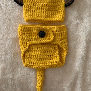 May include: A crocheted lion costume for a baby, featuring a yellow hat with brown ears and a matching yellow nappy cover with a tail.