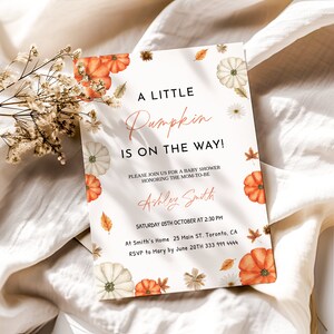 May include: A baby shower invitation with the text "A Little Pumpkin Is On The Way!" in a stylish font. The invitation features watercolor pumpkins, leaves, and floral accents in orange, white, and beige. The invitation includes the name Ashley Smith and event details.