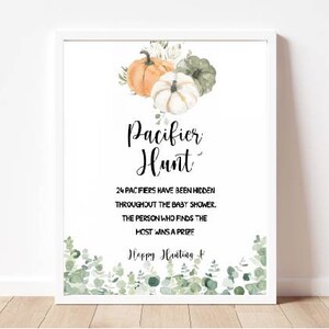 May include: White framed sign with the text "Pacifier Hunt" and details for a baby shower game. The sign features watercolor pumpkins and greenery. The sign is displayed on a wooden surface.