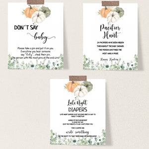 May include: Three baby shower game signs with watercolor pumpkins and greenery. The signs read "Don't Say Baby", "Pacifier Hunt", and "Late Night Diapers". Each sign is taped to the wall with decorative tape.