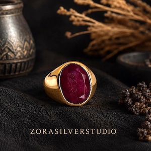 Puede incluir: Un anillo de oro con una gran piedra preciosa de color rojo intenso, tallada en forma ovalada y engastada en una banda de oro lisa y pulida. El texto ZORASILVERSTUDIO está impreso en blanco en la parte inferior, centrado bajo el anillo.