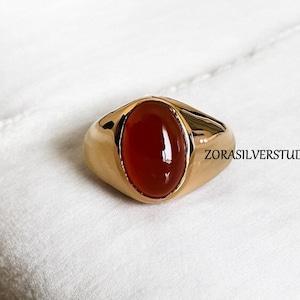 Op de afbeelding: Een gouden ring met een rode ovale edelsteen. De ring ligt op een witte achtergrond.