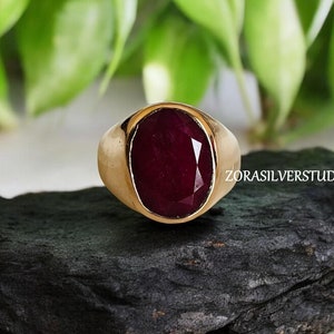 Peut inclure: Une bague dorée ornée d'une grosse pierre précieuse ovale rouge foncé. La bague est posée sur une surface sombre et texturée, avec un feuillage vert en arrière-plan. Le texte "ZORASILVERSTUDIO" est visible sur le côté droit de la bague.