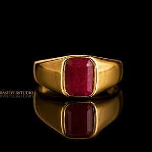 Könnte beinhalten: Ein goldener Ring mit einem rechteckigen, tiefroten Edelstein. Der Ring hat eine polierte, glatte Oberfläche und ist vor einem schwarzen Hintergrund platziert. Die Reflexion des Rings ist darunter sichtbar.