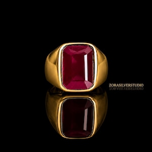 Può includere: Un anello d'oro con una grande gemma rettangolare rosso scuro. L'anello è posto su uno sfondo nero, con un riflesso dell'anello visibile sotto. L'anello è uno stile a sigillo.