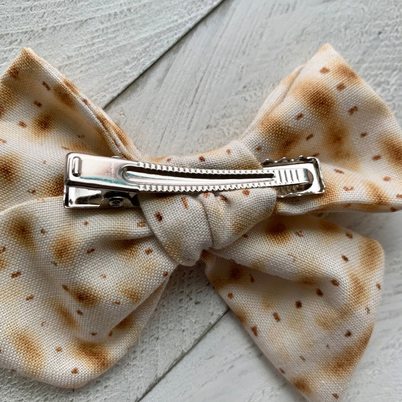 May include: A white fabric bow with a brown matzah pattern and a silver alligator clip.