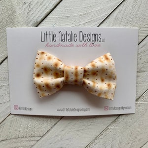 May include: A bow tie made of fabric with a matzah pattern. The bow tie is on a white card with the text "Little Natalie Designs LLC handmade with love".