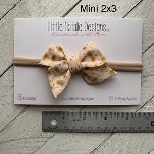 May include: A small, beige bow with a brown speckled pattern, tied on a nude elastic headband. The bow measures 2 inches by 3 inches. The headband is handmade with love.