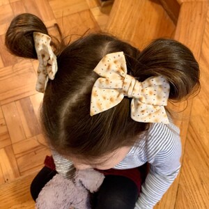 May include: A young girl with brown hair wearing two brown and white patterned hair bows. The bows are tied in a bow shape and are placed on top of two messy buns.