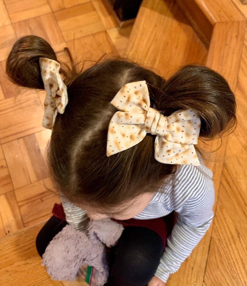 May include: A young girl with brown hair wearing two brown and white patterned hair bows. The bows are tied in a bow shape and are placed on top of two messy buns.
