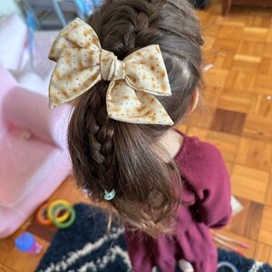 May include: A young girl with brown hair wearing a burgundy dress has a brown and white bow in her hair. The bow is made of fabric with a pattern that resembles matzah.