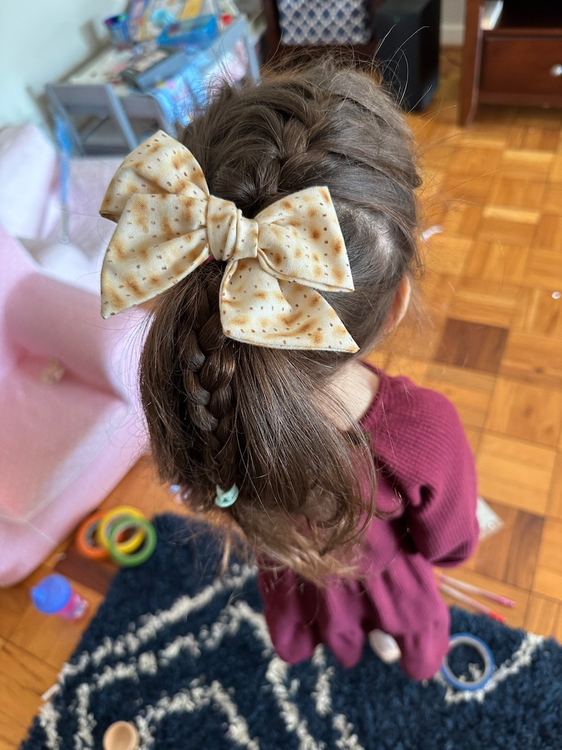 May include: A young girl with brown hair wearing a burgundy dress has a brown and white bow in her hair. The bow is made of fabric with a pattern that resembles matzah.